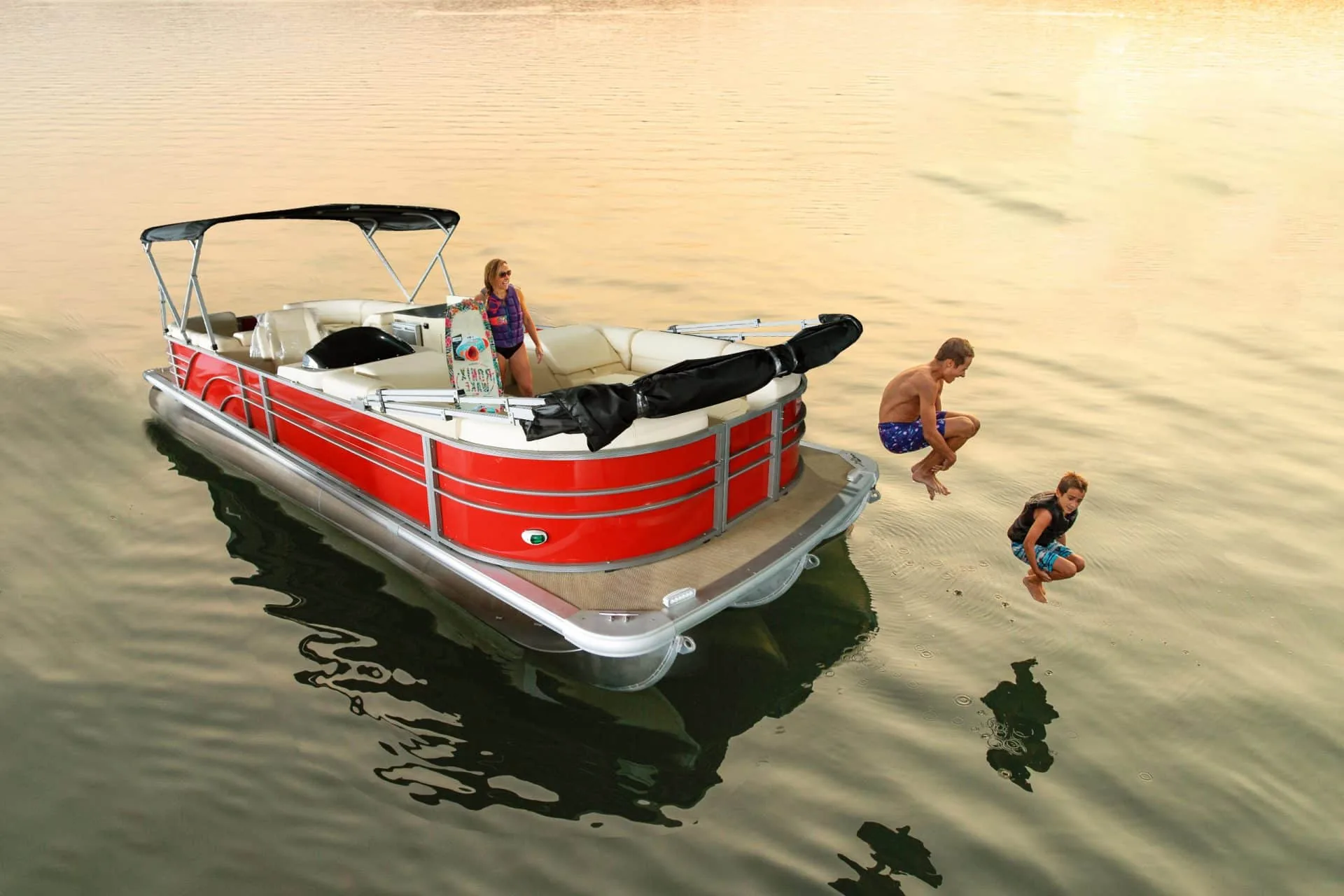 Red pontoon boat with a bimini frame, a passenger aboard and swimmers alongside